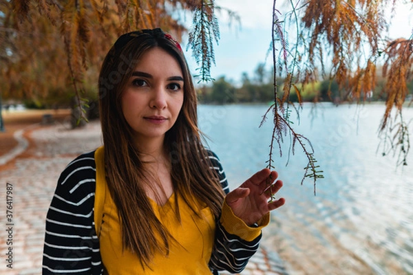 Fototapeta A young woman at autumnal park beside a lake looking at camera while touching a branch