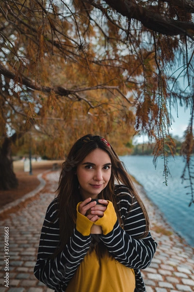 Fototapeta A pretty caucasian woman holding a cup of coffee to go at autumnal park. Vertical, copy space