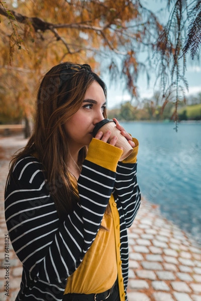 Fototapeta A young caucasian woman drinking coffee at autumnal park while looking at lake. Vertical