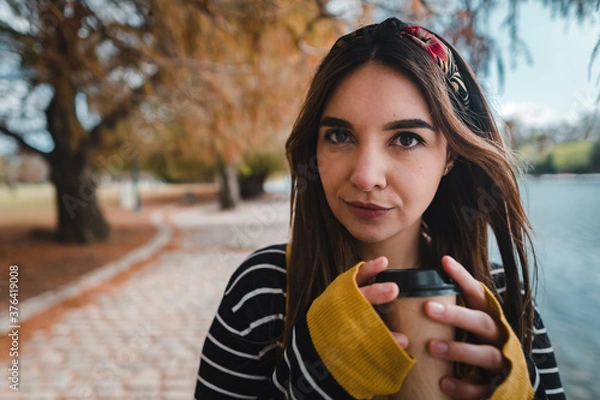 Fototapeta A pretty caucasian woman holding a cup of coffee and looking at camera at autumnal park
