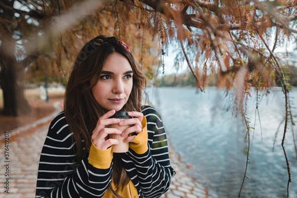 Fototapeta A pretty caucasian woman holding a coffee while standing below autumnal tree beside lake