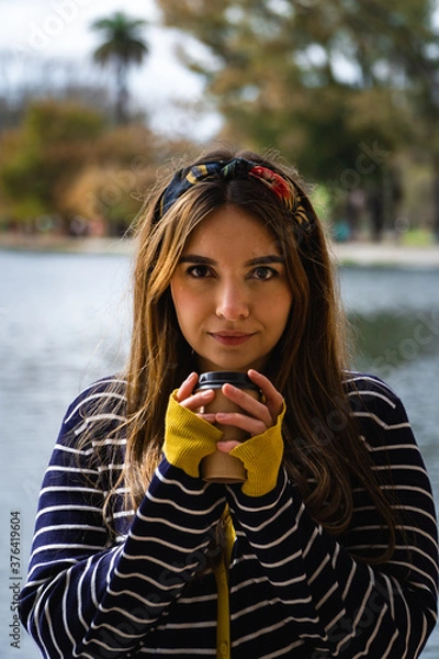 Fototapeta A pretty caucasian woman holding a cup of coffee while standing against a lake at park