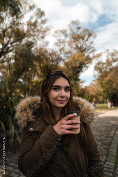 Fototapeta A pretty caucasian woman smiling while holding a coffee and enjoying a walk at park. Vertical