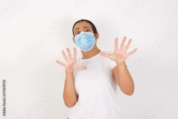 Fototapeta Young arab woman wearing medical mask standing over isolated white background afraid and terrified with fear expression stop gesture with hands, shouting in shock. Panic concept.
