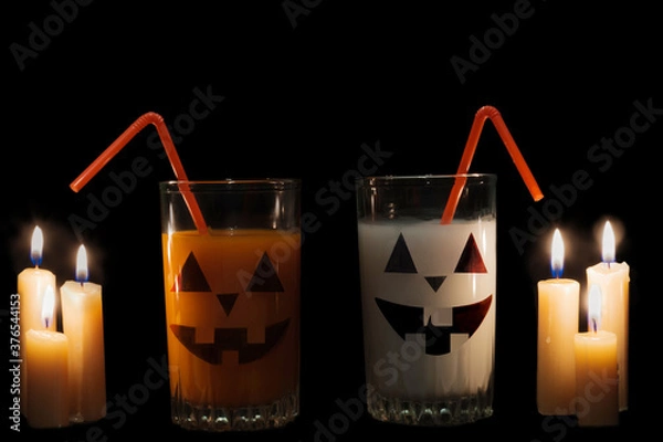 Obraz There are two glasses of white and orange drinks on a black background, lit by candles, and on the glasses are an eerie smiling face. Halloween. Idea - drinks for the holiday