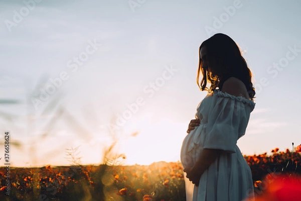Fototapeta Silhouette of beautiful pregnant woman standing on poppies field in sunset. Pregnant woman in white dress relaxing outdoors. Copy space.