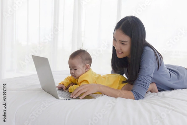 Fototapeta An Asian mom is working on a laptop in bed and with her baby next to the concept of work at home.