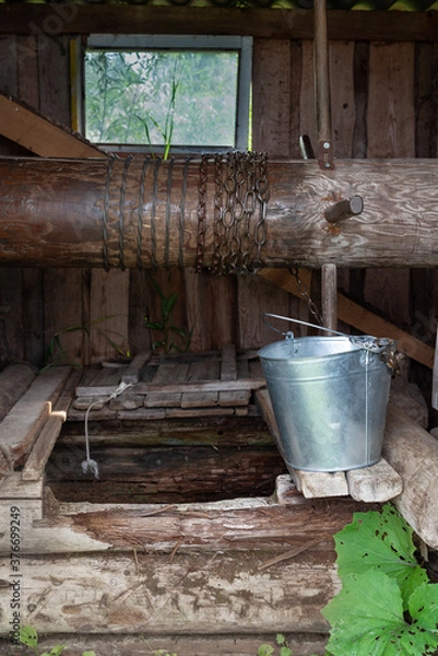 Obraz An old village well, with a metal bucket, on a summer day, in Russia. Close-up, vertical format.