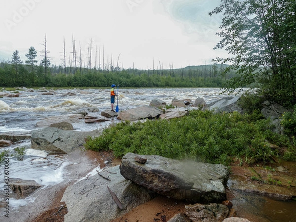 Fototapeta a boy with a paddle in a helmet with a life jacket standing on a large rock near a mountain river