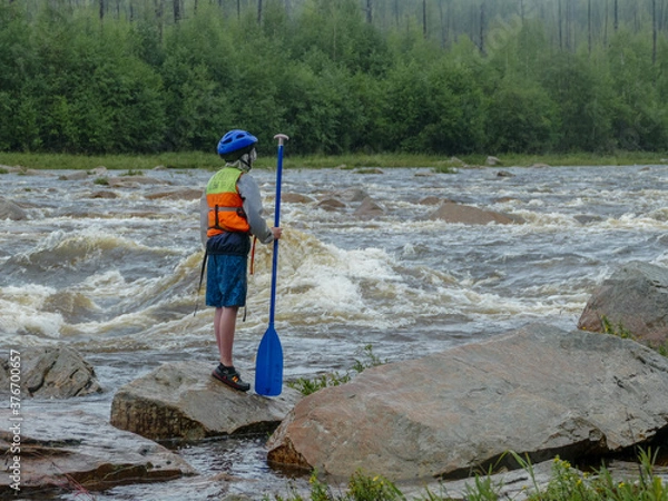 Fototapeta a boy with a paddle in a helmet with a life jacket standing on a large rock near a mountain river