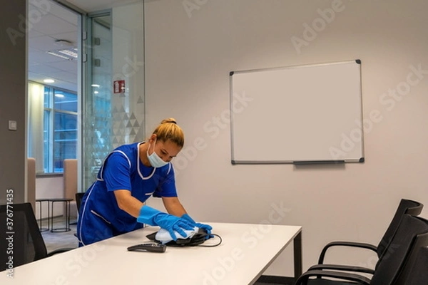 Fototapeta A cleaning lady with a mask on her face cleans the meeting room.