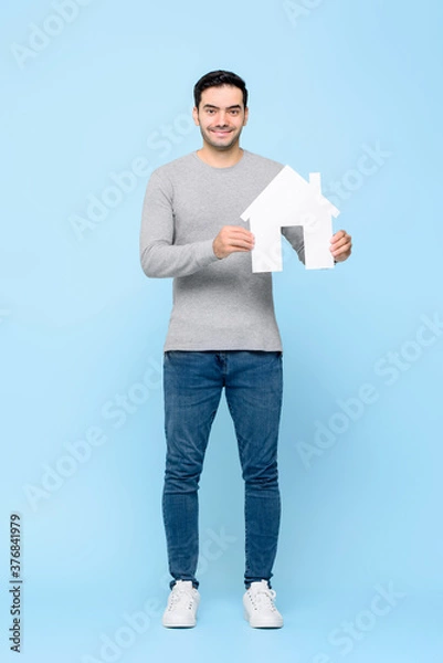 Fototapeta Full length portrait of smiling cheerful Caucasian man holding house model cutout standing in light blue isolated studio background