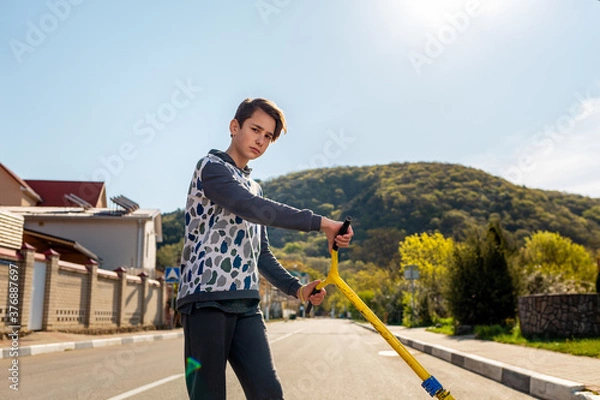 Fototapeta A confident teenage boy with an arrogant look and a scooter in his hand.In the background,a street and road on a Sunny day,a view of the mountain.Copy space.Concept of sports and adolescent activities