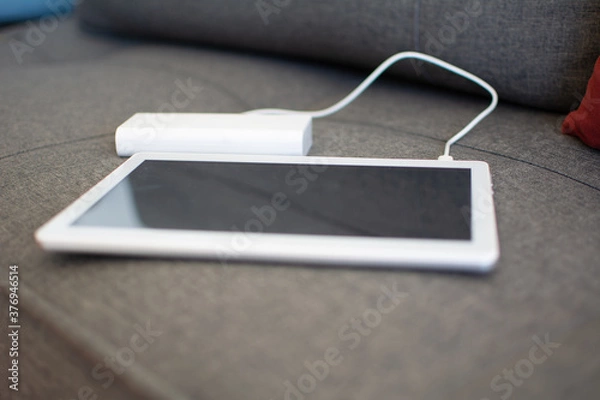 Fototapeta A white tablet charging energy from a power bank on the sofa. blur background with copy space