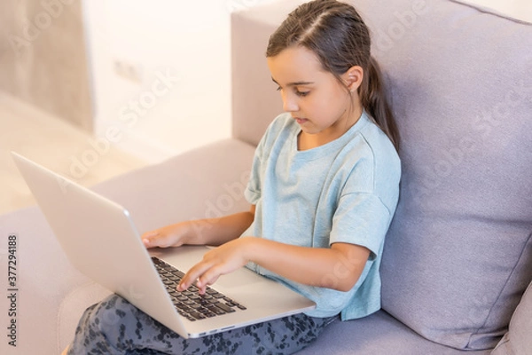 Fototapeta Little girl doing her homework at home and using a laptop