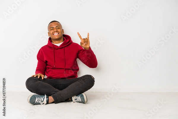 Fototapeta Young latin man sitting on the floor isolated showing a horns gesture as a revolution concept.