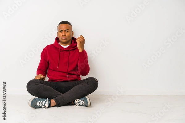 Fototapeta Young latin man sitting on the floor isolated showing fist to camera, aggressive facial expression.