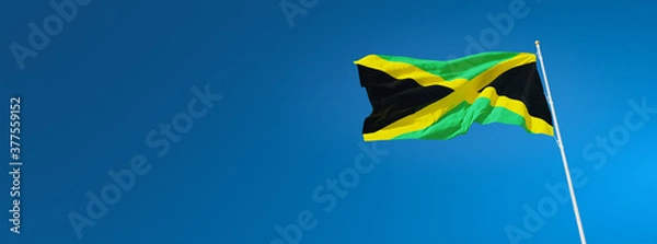 Fototapeta The beautiful flag of Jamaica waving against a blue sky in wide format. Jamaican national flag on background