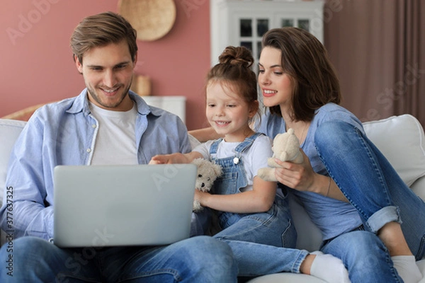 Obraz Positive friendly young parents with smiling little daughter sitting on sofa together answering video call on laptop and waving hand in greeting while relaxing at home on weekend