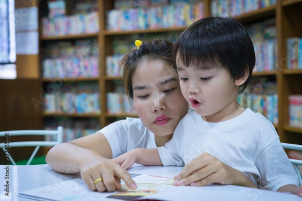 Fototapeta Asian young mother with little boy reading book together in library. Happy family. Parent educating children. Pre-school learning concept.