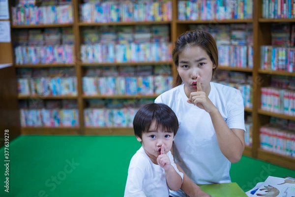 Fototapeta Asian young mother with little boy reading book and holding finger on lips making a silent gesture together in library. Parent educating children. Pre-school learning concept.