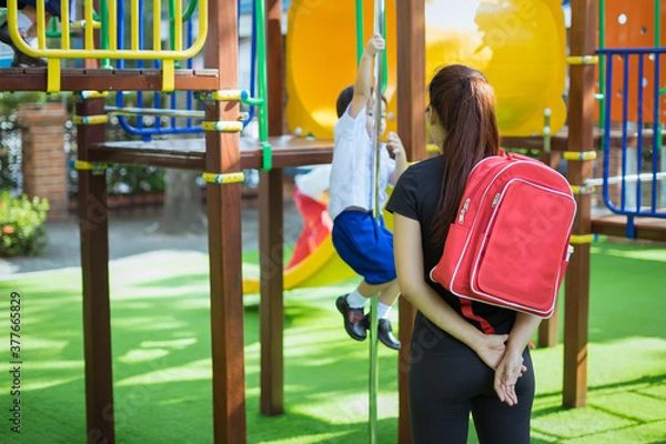Fototapeta Mother with backpack behind the back watching her son playing around in the playground. Education, childhood, family and people concept.