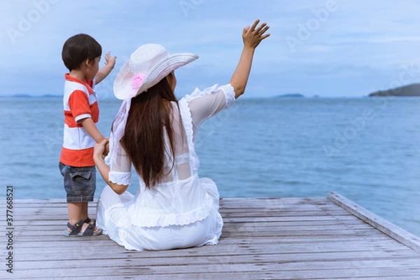 Fototapeta Young mother in long dress and son waving goodbye on wooden bridge, on a background of the sea.