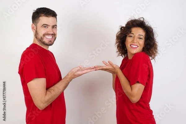 Fototapeta Young beautiful couple wearing red t-shirt on white background pointing aside with hands open palms showing copy space, presenting advertisement smiling excited happy
