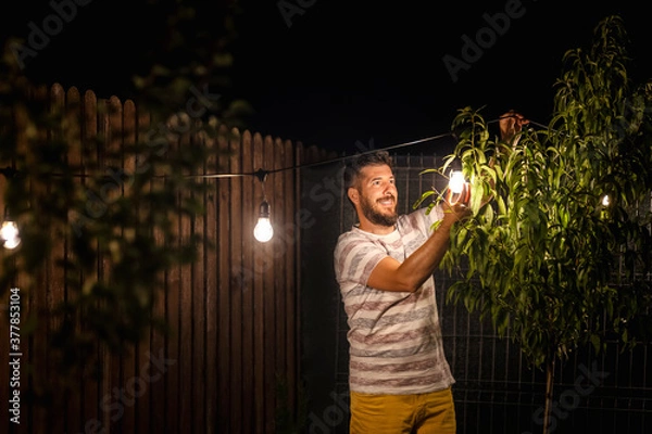 Fototapeta Party time in backyard with happy man hanging string lights in trees