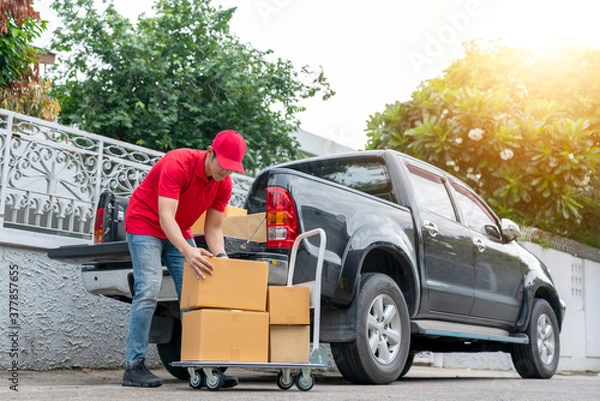 Obraz Delivery men in red uniform unloading cardboard boxes from pickup truck. Courier man sending the parcel or package to the customer on a business day. Online shopping and transport logistics concept.