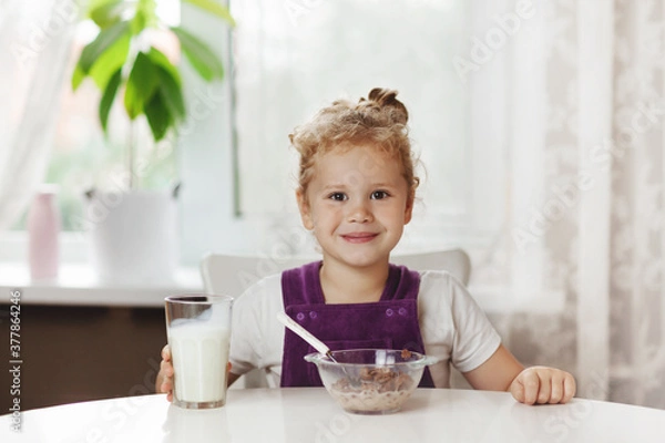 Fototapeta Charming girl eats cereal with milk for Breakfast and . On the table are chocolate flakes, a glass of milk. Healthy Breakfast, taking care of children. Space for text