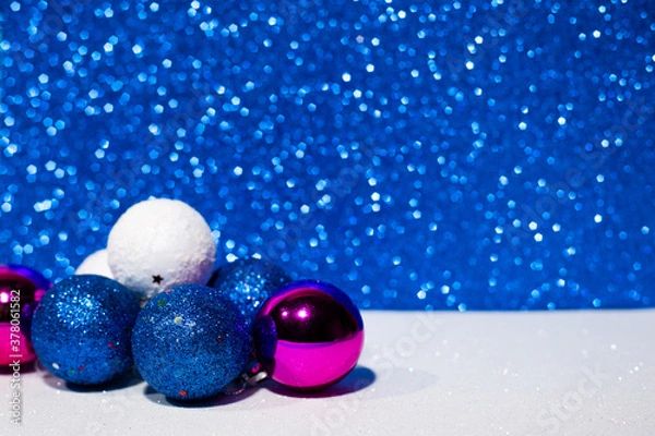 Fototapeta Decorative Christmas balls in blue, white and purple on the left on a white surface. Blue Christmas lights on the background. Side view.Place for inscriptions and greetings