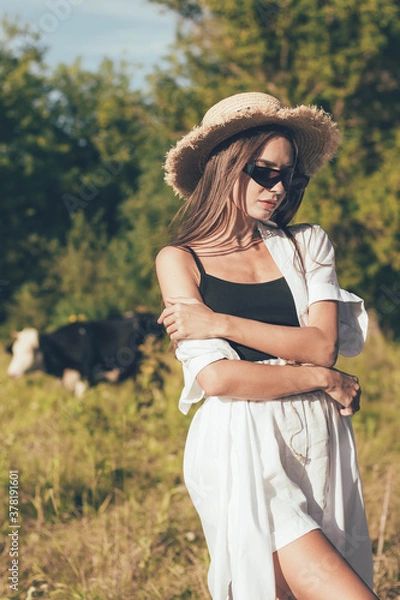Fototapeta A woman in stylish glasses in a suit and a hat stands in a field near a cow