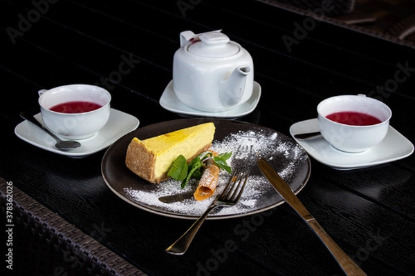 Obraz Close-up on a black table is a tea set with fruit tea, a black plate with cheesecake, mint and cutlery.