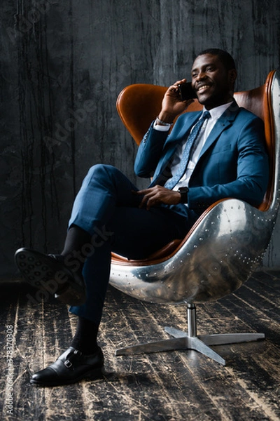 Fototapeta A black man in a blue suit sits in a loft aviator chair, talking on the phone and smiling