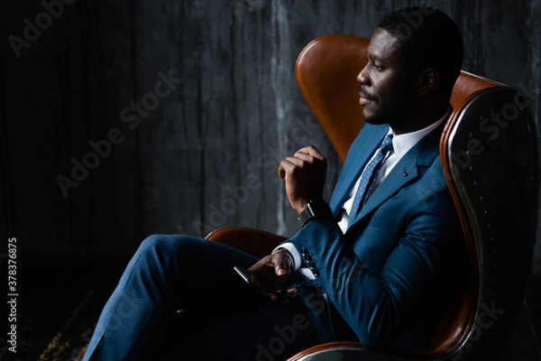 Obraz African American businessman in a blue classic suit sits in an aviator loft armchair with a phone in his hands
