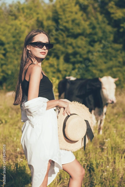 Fototapeta 
A slender woman of model appearance in stylish glasses holds a hat in her hands and is in nature near a cow