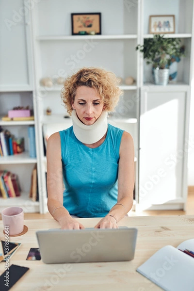Fototapeta Woman with neck / cervical collar and neck / spinal injury working at home on a laptop.