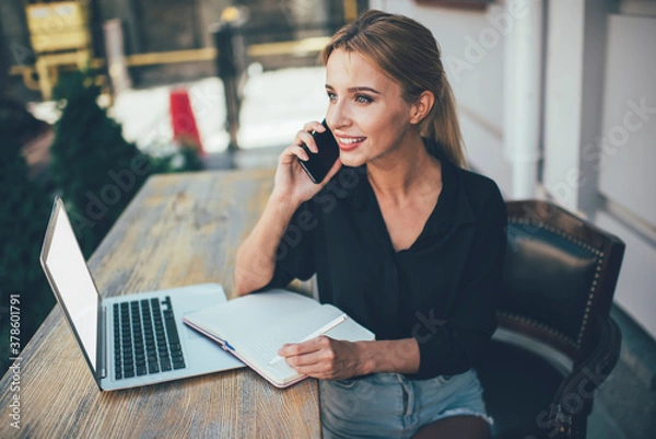 Fototapeta Happy female student receiving positive cellphone call sitting outdoors with laptop device and education textbook for e learning, prosperous freelancer communicate via smartphone and smiling