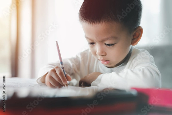 Fototapeta Asian Boy Doing homework with the intention. Child boy holding pencil writing, A boy drawing on white paper at the table,Elementary school and home schooling, Distance Education concept.