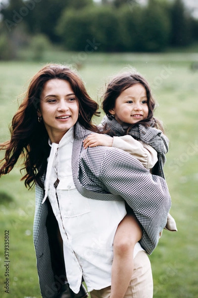 Obraz Stylish mother and daughter have fun outdoors in a field with green grass by the river when it rains