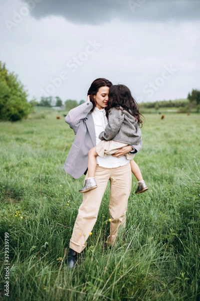Obraz Stylish mother and daughter have fun outdoors in a field with green grass by the river when it rains
