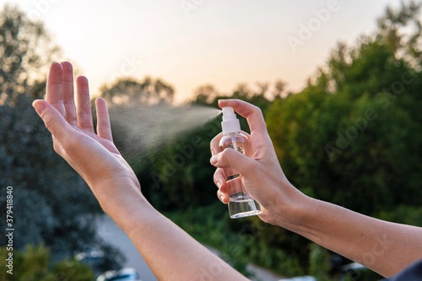 Obraz Woman use an alcohol hand sanitizer from a spray bottle to clean her hands and palms for protection from viruses, bacteria, and coronavirus. Beautiful golden sky in the background and green trees.