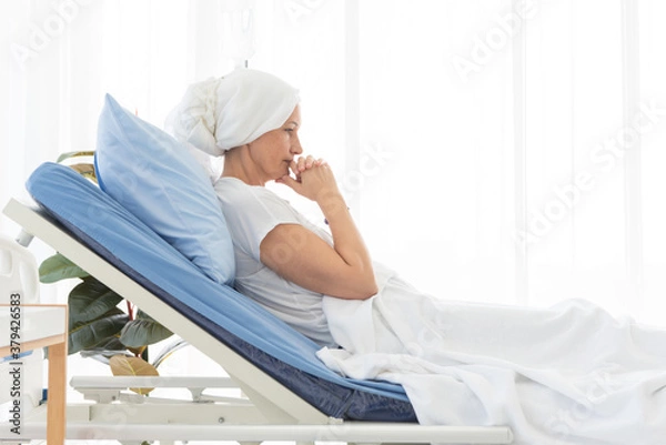 Obraz Side view of senior breast cancer patient wearing a headscarf laying in bed and praying in the hospital room, cancer concept