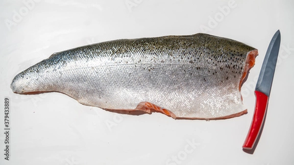 Fototapeta 
Salmon fillet with silver scales dotted with spots in the shape of small black circles, next to a red kitchen knife, on a white background