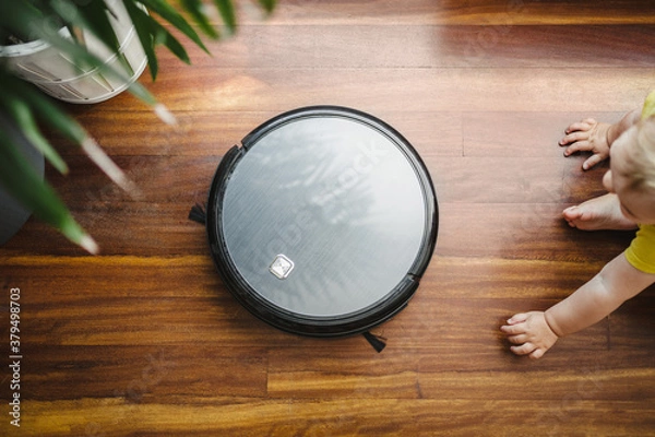 Fototapeta a robot vacuum cleaner cleaning the floor with a baby dressed in yellow and a plant