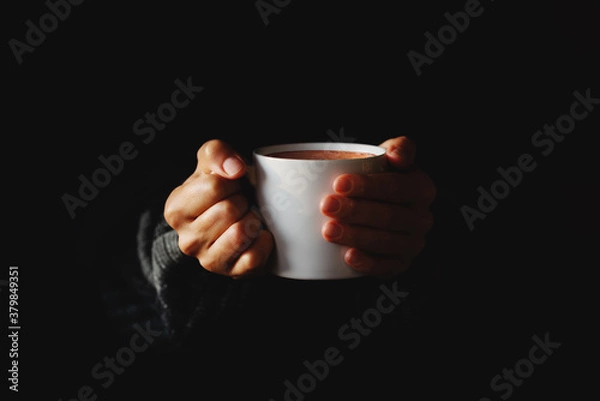 Fototapeta Cup of coffee in women's hands on black background. Woman warming her hands on mug of hot beverage in the dark. Copy space. Minimal style. Low key.