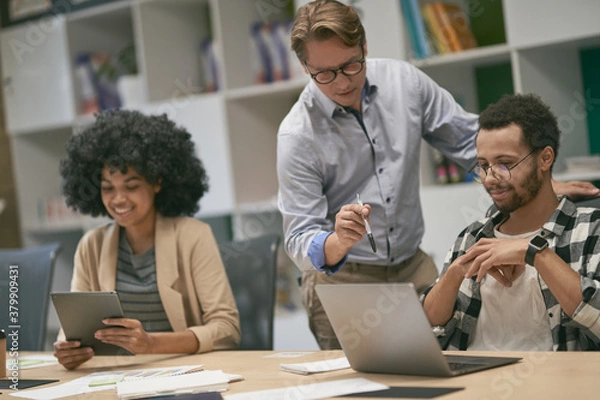 Fototapeta Confident caucasian male boss looking at the laptop screen and discussing project results with two young diverse colleagues sitting at the desk