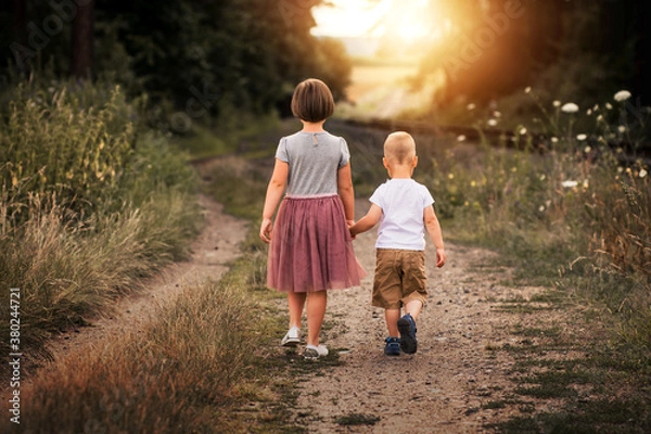Fototapeta Two siblings walking in nature in the sunset. Brother and sister go on path and holding hands. Back view.