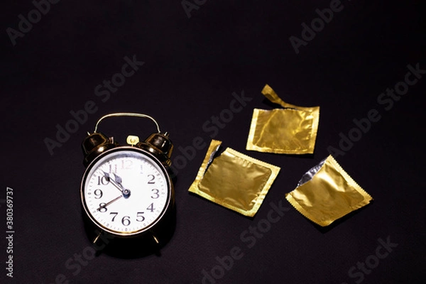Fototapeta A gold watch is located on a black background next to three opened condoms. Special design for the contraception day. There is a space for text.